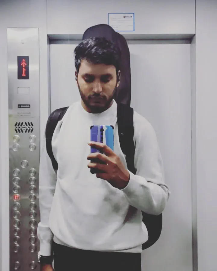 A man in a white sweatshirt photographing himself in a lift mirror, a guitar case slung across his back.