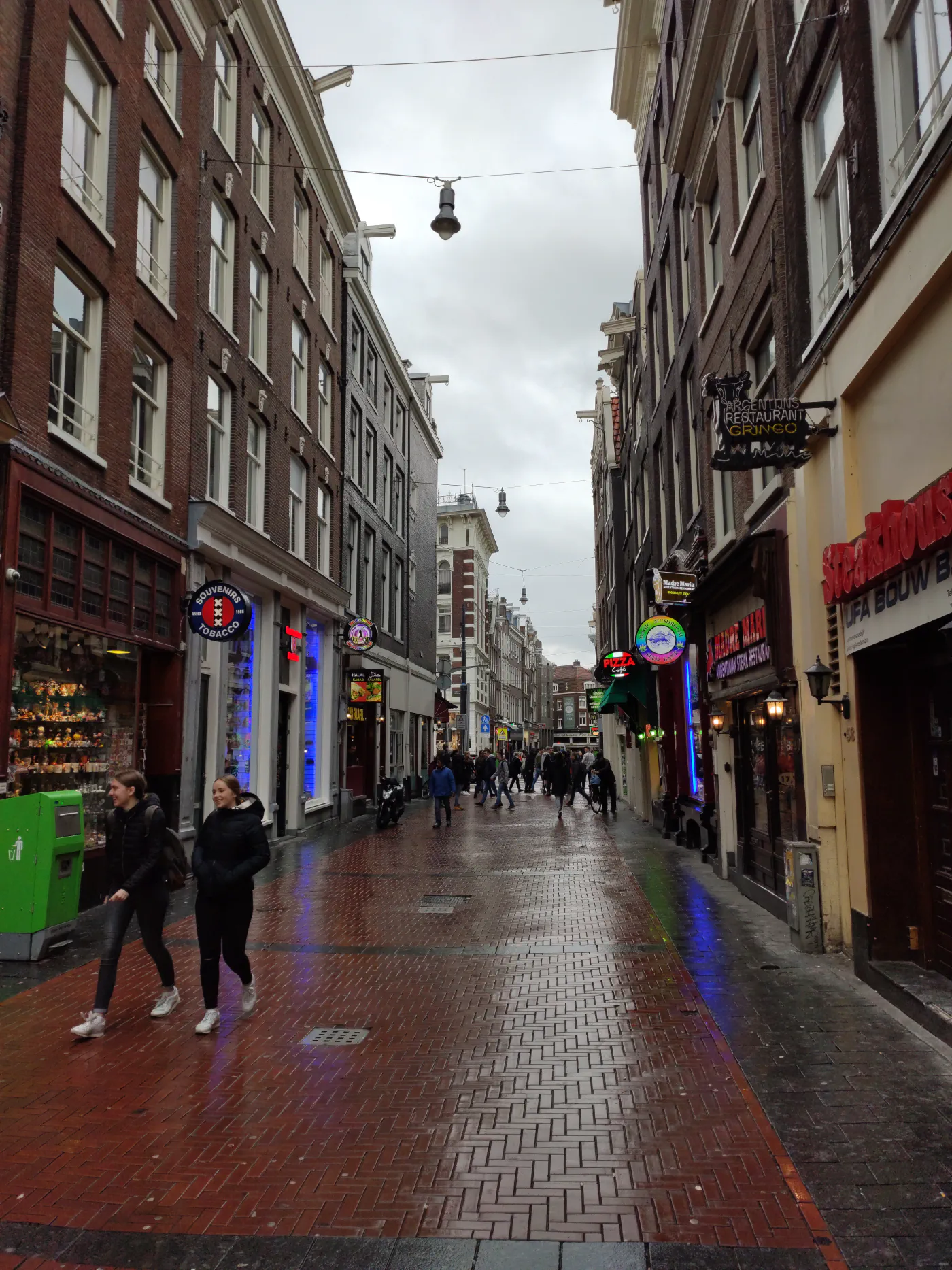 A narrow Amsterdam street of tall brick buildings on a grey day, neon signs for restaurants and cafés along the ground floor, people walking on wet brick paving.