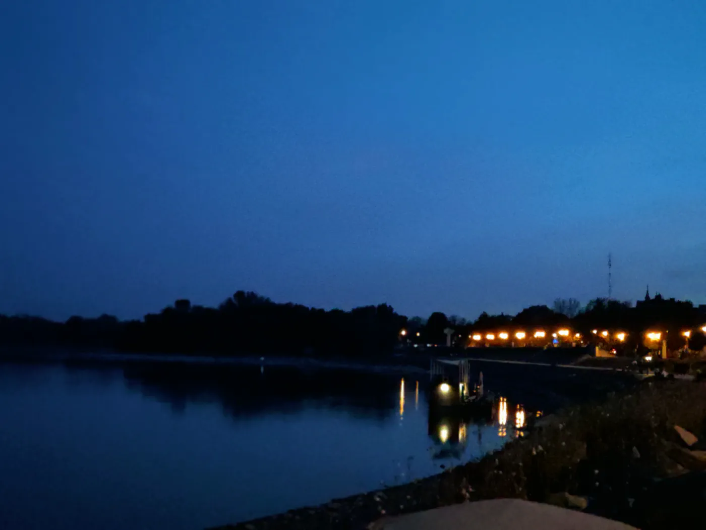 The Danube at blue hour, still water reflecting a line of warm street lamps along the far bank, dark treeline and a church spire on the horizon.