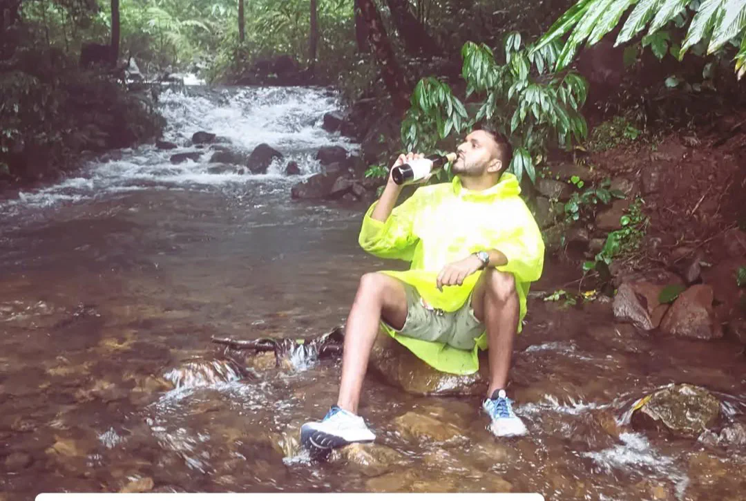 A man in a bright yellow rain poncho sitting on a rock in the middle of a fast-flowing forest stream, drinking from a bottle, dense wet green forest all around.
