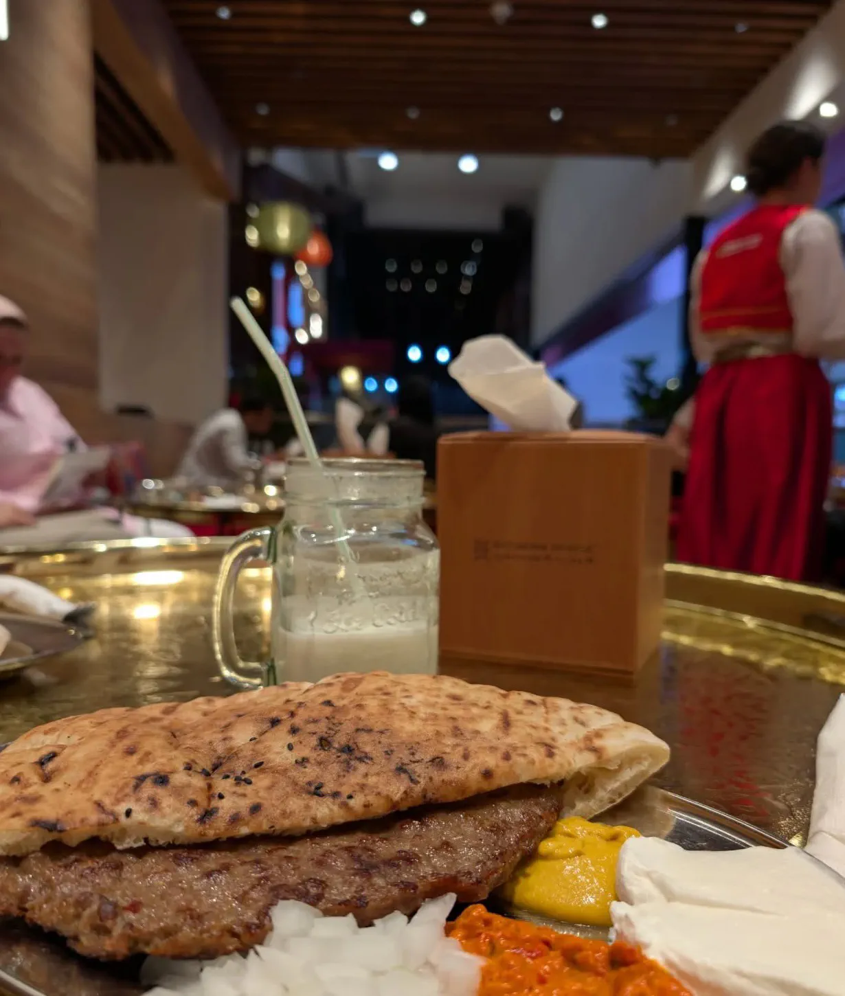 A metal platter holding grilled ćevapi sausages tucked under a folded flatbread, with chopped raw onion, red ajvar, mustard and white kajmak alongside, in a warmly lit restaurant.