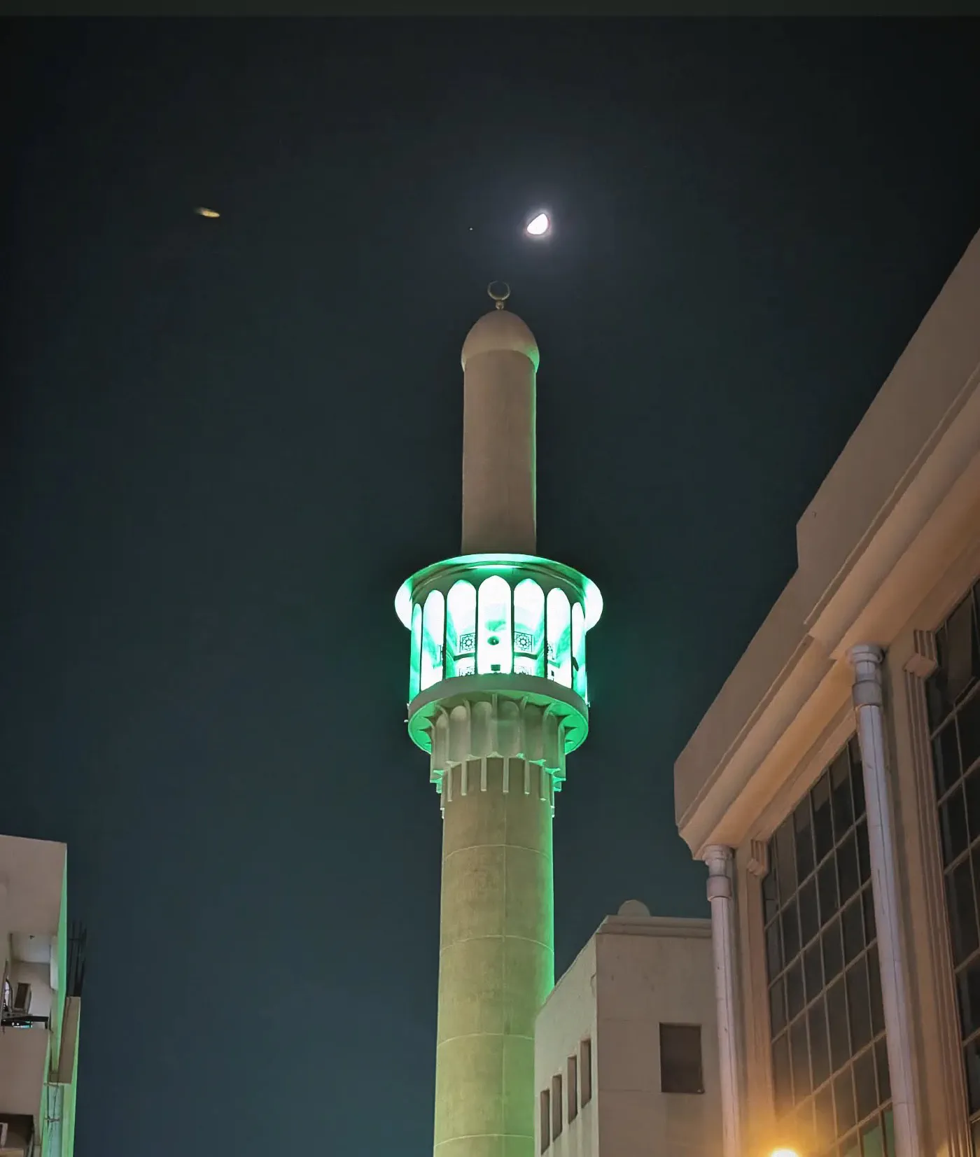 A slender minaret lit green against a dark blue night sky, a crescent moon directly above it, older low buildings on either side.