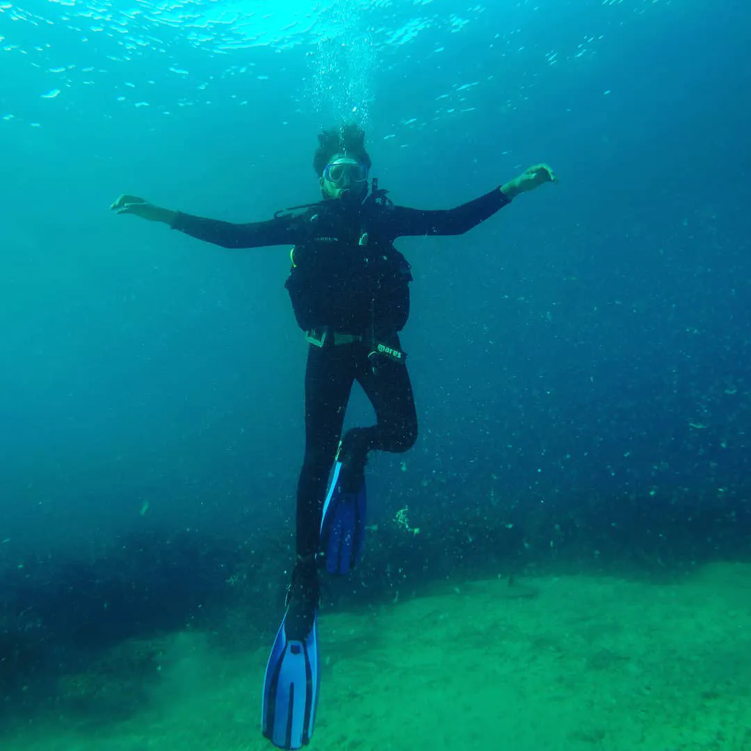 A diver in a black wetsuit suspended mid-water in clear blue sea, arms outstretched, bubbles rising toward the surface above.