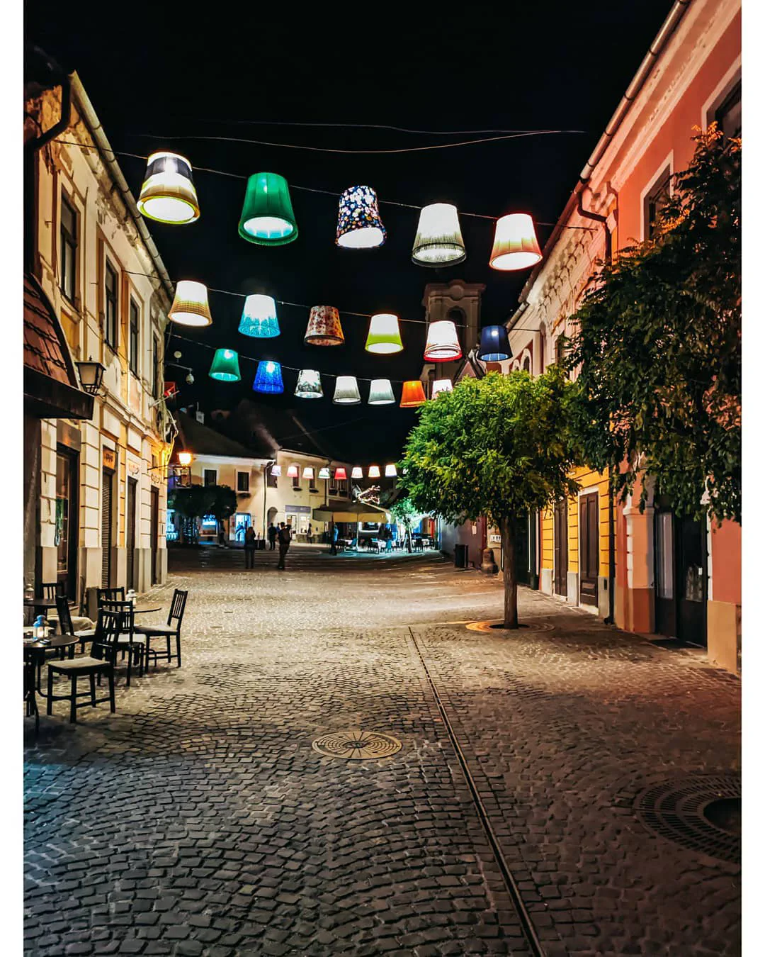 A cobbled street at night strung with rows of mismatched, brightly coloured lampshades glowing above empty café tables.
