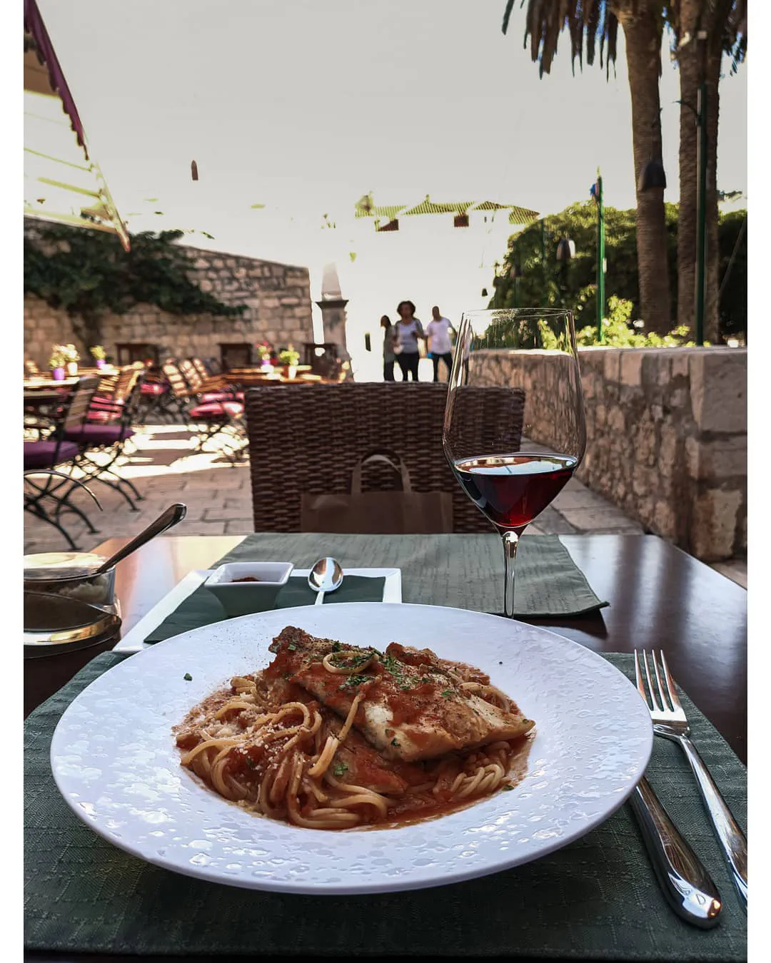 A plate of spaghetti with fish in tomato sauce and a glass of red wine on a terrace table, with stone walls, palm trees and bright sky beyond.