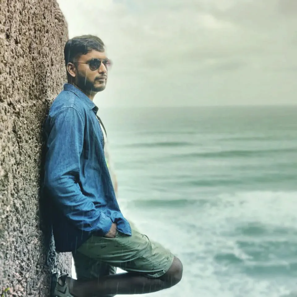 A man in a denim shirt and sunglasses leaning against a rough stone wall above a rough green sea, rain falling.