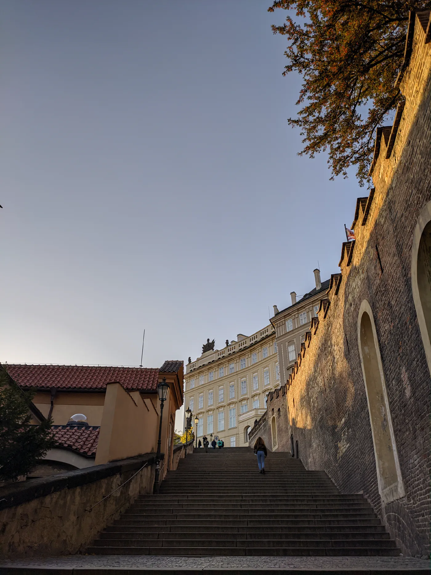 Stone steps climbing toward Prague Castle in low golden evening light, a high brick wall on the right, an autumn tree overhanging, a lone figure walking up.