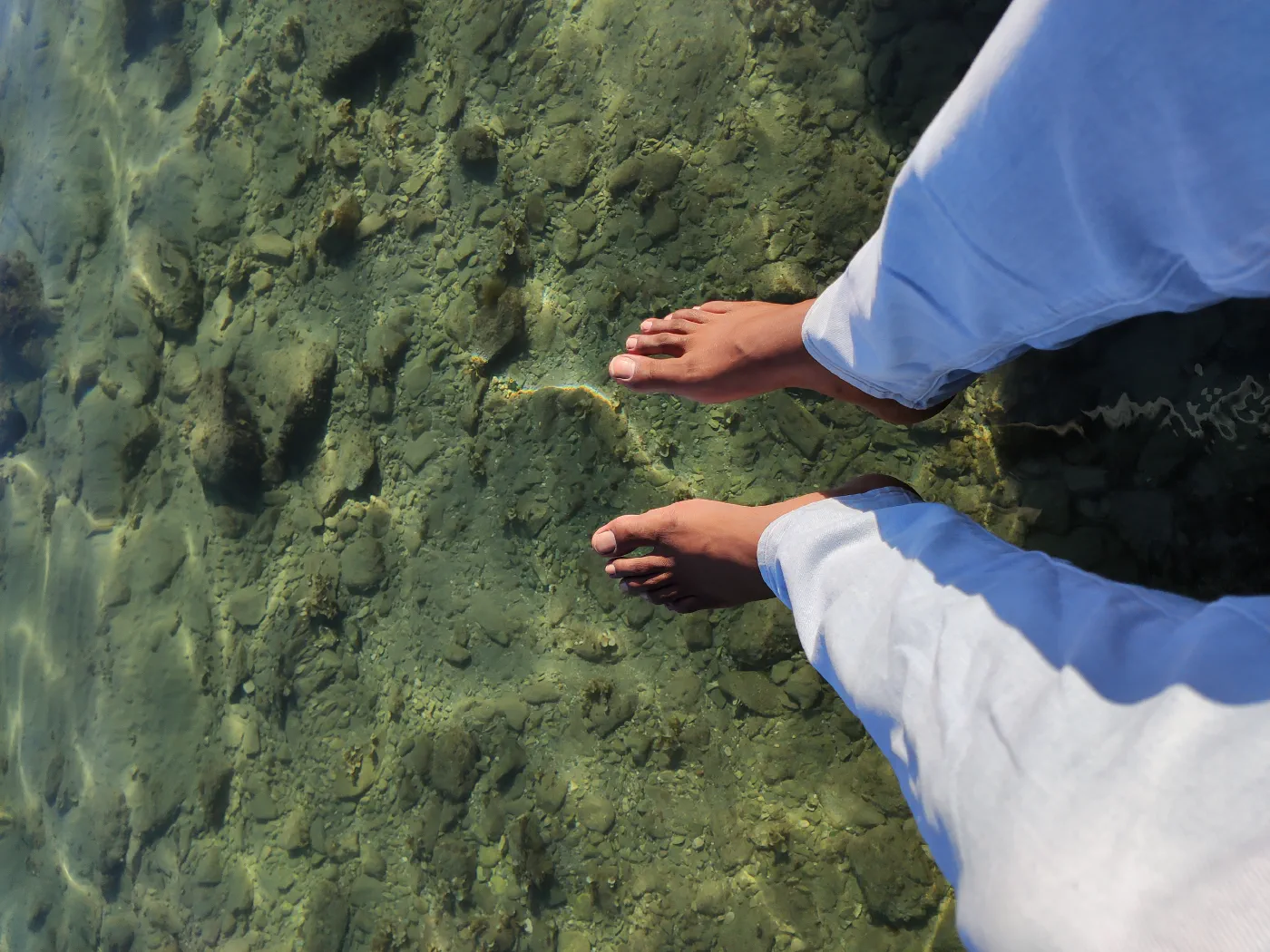 Looking down at bare feet standing in shallow, perfectly clear turquoise sea over pale round pebbles, white trousers rolled up.