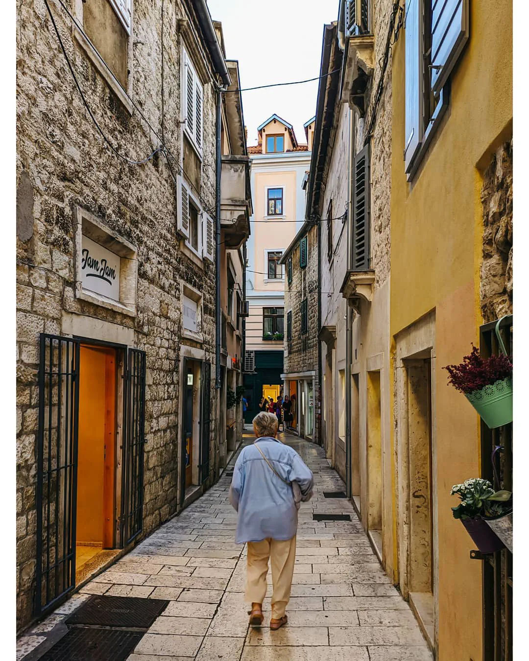 A narrow stone alley in Split's old town, washed in warm light, an elderly woman walking away from the camera between shuttered buildings.