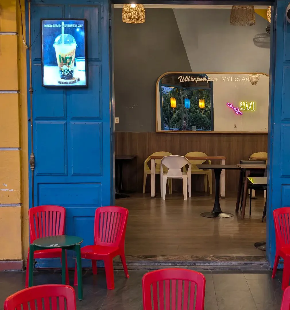 A café front in Hoi An with an ochre-yellow wall and bright blue folding doors, a neon sign reading IVY coffee-tea, a yellow silk lantern above, and red plastic chairs around small tables on the pavement.